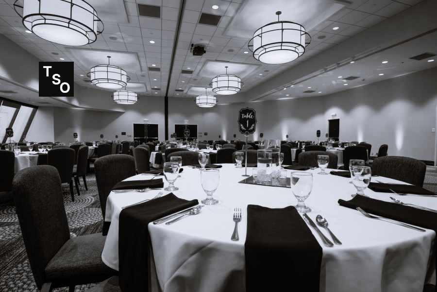 Sala grande con mesas preparadas para una comida y al fondo pantallas para oradores, representando un evento corporativo